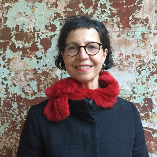 Photo of Rita Orsini, she wears round tortise shell glasses, a short black fringe and bob, silver earrings, a red lipstick smile and a black coat. She stands in front of a textured wall that has peeling paint with orange, aqua and white colours. It is a head and shoulders shot